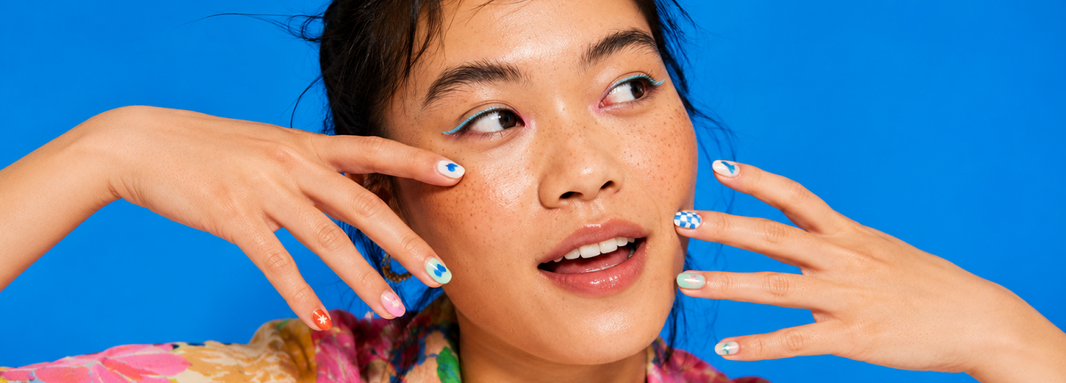 A close-up lifestyle shot of a woman with freckles and blue eyeshadow, touching her face with manicured nails against a solid bright blue background.