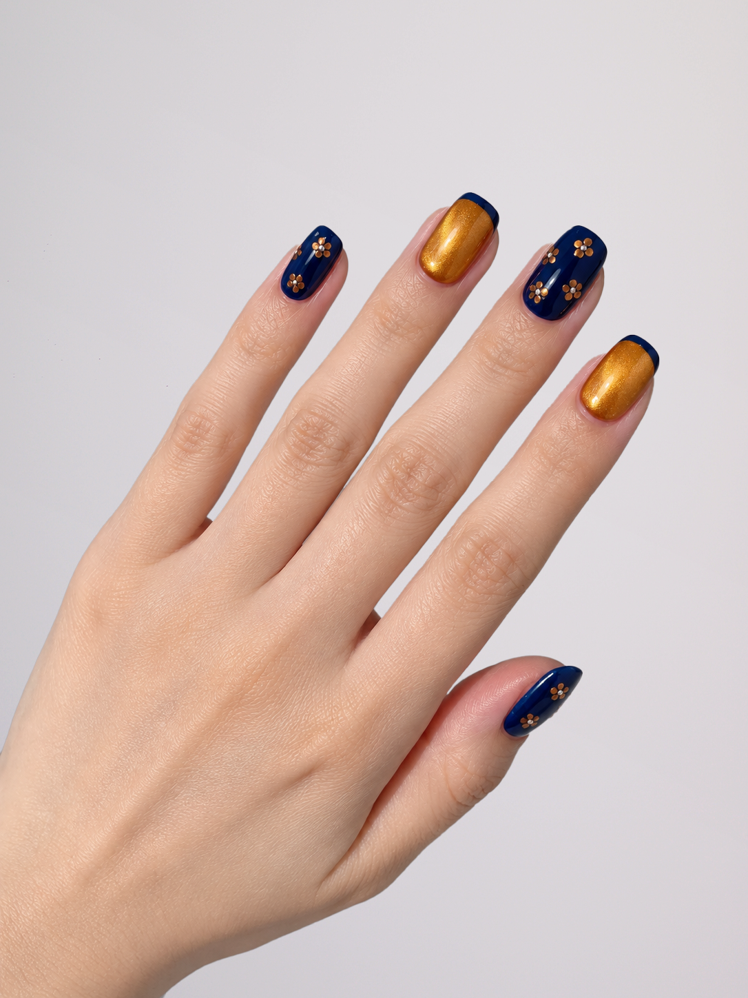 A close-up of a hand displaying manicured nails with a navy blue and metallic gold color scheme. Two nails feature a solid metallic gold finish, while the other two are navy blue with small gold star patterns.