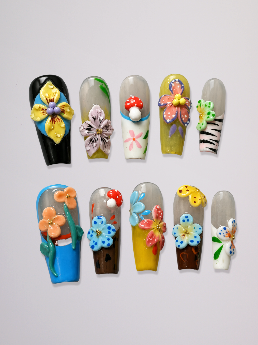 Ten coffin-shaped press-on nails arranged in two rows on a light gray background. The nails feature various colorful 3D floral, mushroom, zebra, and landscape designs on clear or colored bases.
