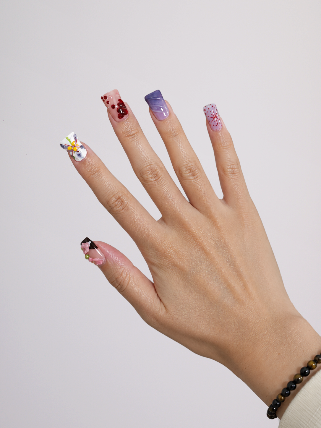 A close-up of a hand displaying a variety of colorful nail art designs, including glitter, floral patterns, and abstract gradients, set against a plain light gray background.