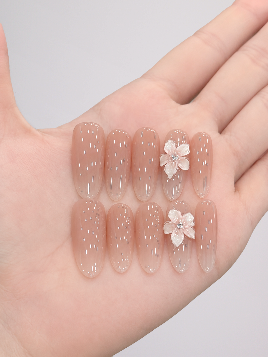 A close-up of a person's hands showcasing a manicure with glossy nude pink glitter nail polish and delicate white flower decals on the nails, set against a solid light gray background.