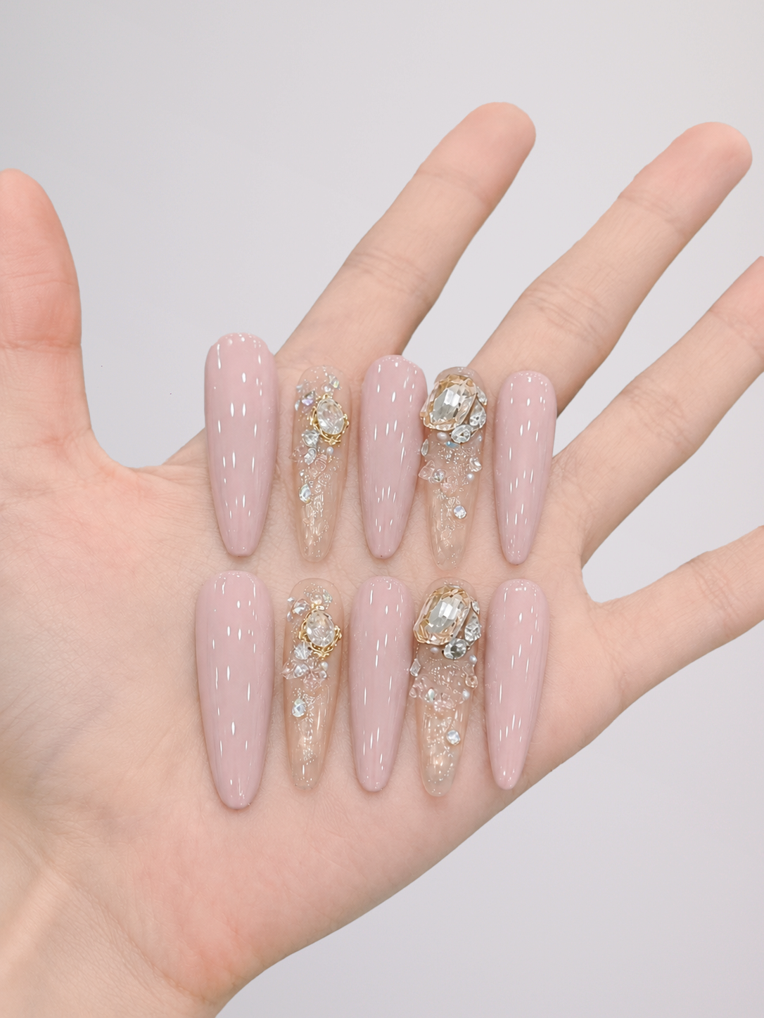 A hand displaying a set of long, almond-shaped press-on nails. The nails feature a glossy pale pink base with two central clear rhinestone-embellished designs and two clear rhinestone-embellished designs on the lower row.
