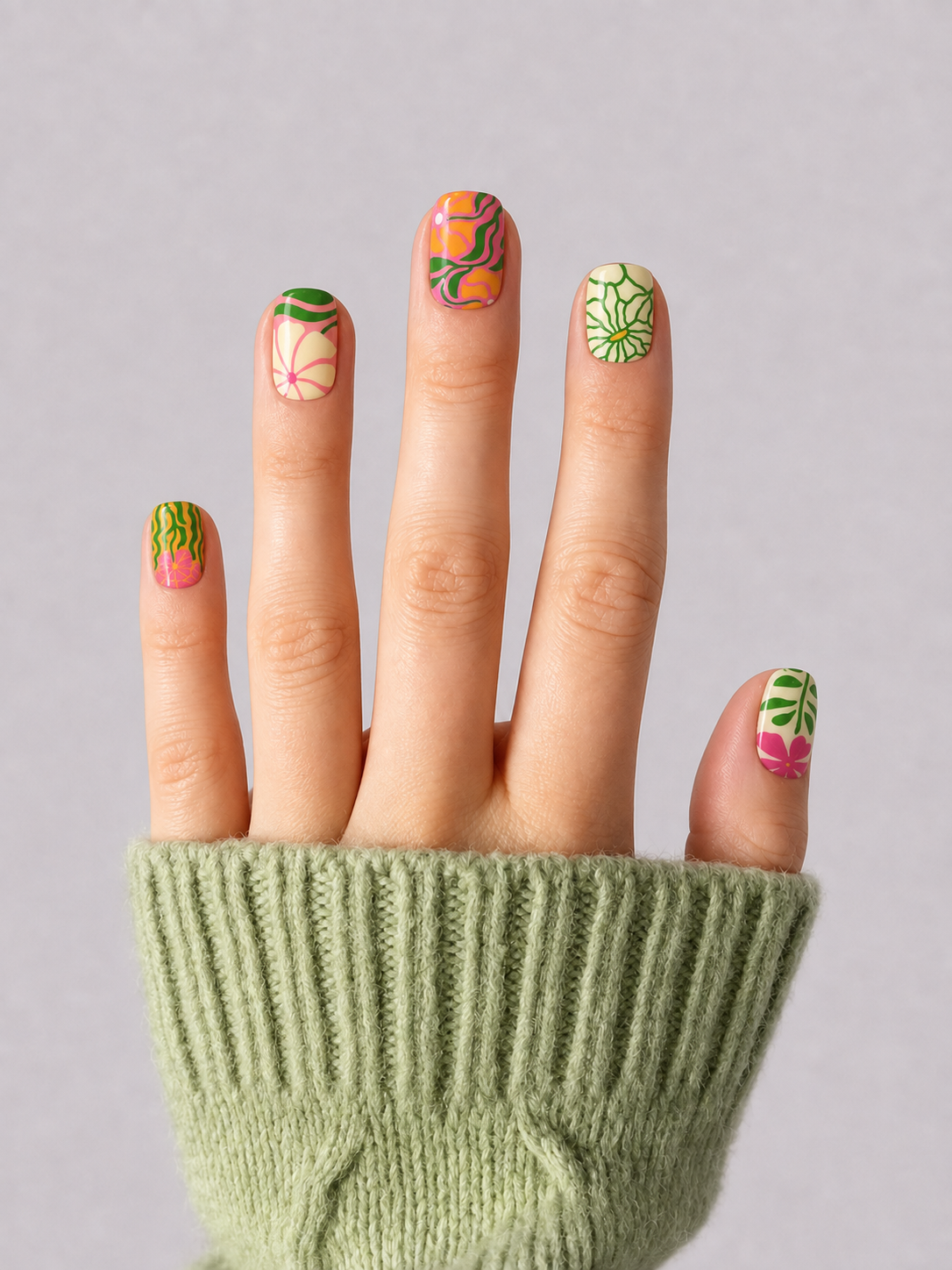 A close-up of a hand wearing a light green ribbed knit sweater, showcasing colorful tropical-themed nail art with orange, pink, and green designs on a neutral gray background.