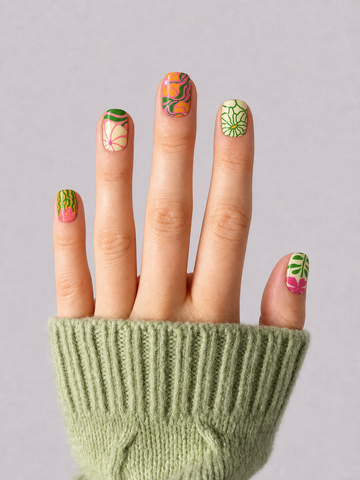 A close-up of a hand wearing a light green ribbed knit sweater, showcasing colorful tropical-themed nail art with orange, pink, and green designs on a neutral gray background.