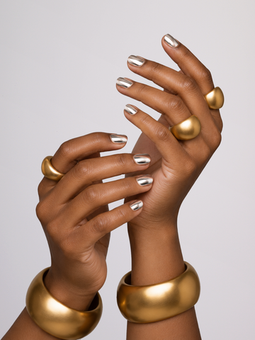 Two hands with metallic silver nail polish wearing chunky gold bangles and matching gold rings, set against a solid light gray background.