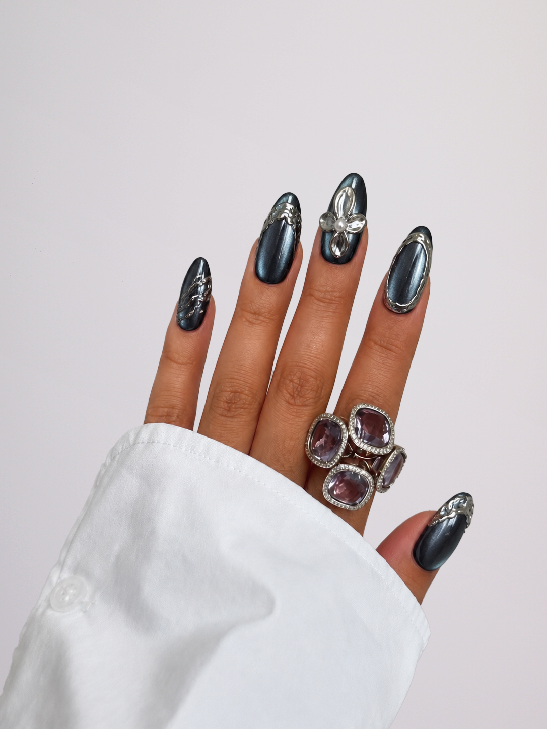 A close-up of a hand with long, glossy black stiletto-shaped nails decorated with silver butterfly and floral details, wearing a chunky silver ring with four oval purple gemstones, styled with a white shirt cuff against a light gray background.