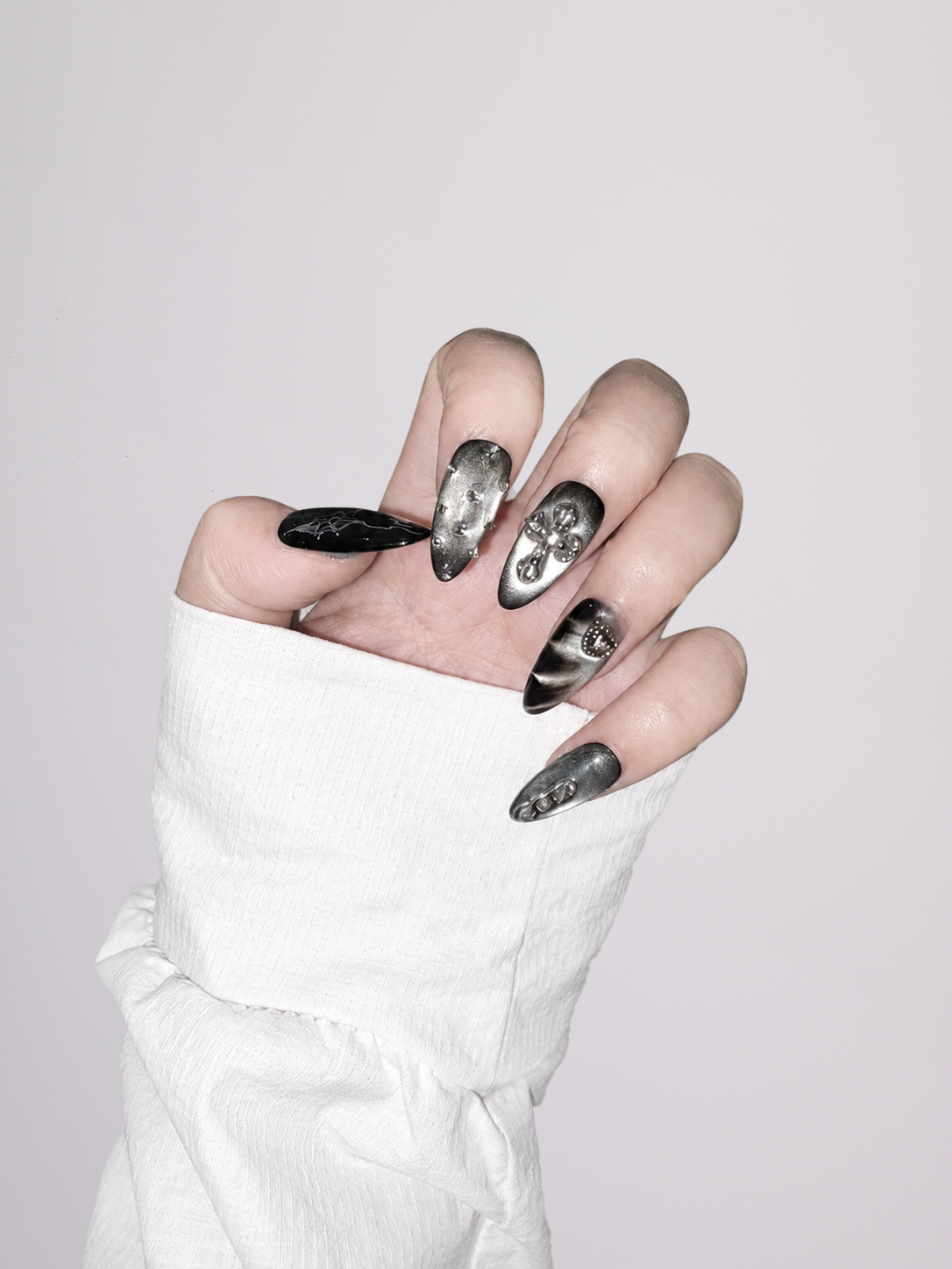 A hand wearing long, glossy black stiletto-shaped nails with silver glitter and skull designs, styled with a white sleeveless top against a light gray background.