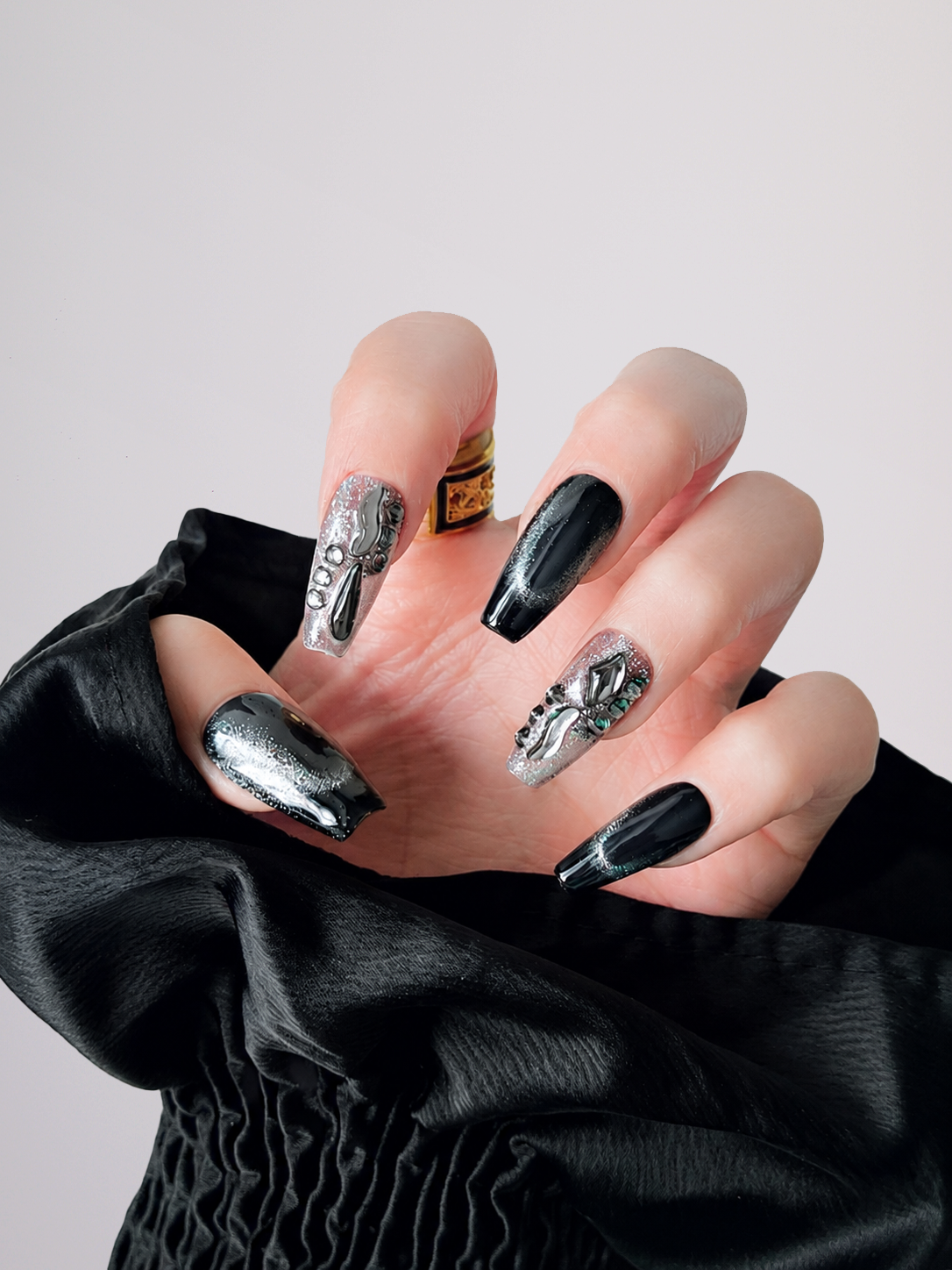 A close-up of a hand with long almond-shaped nails styled in a gothic black and silver manicure, wearing a black draped sleeve against a light gray background.