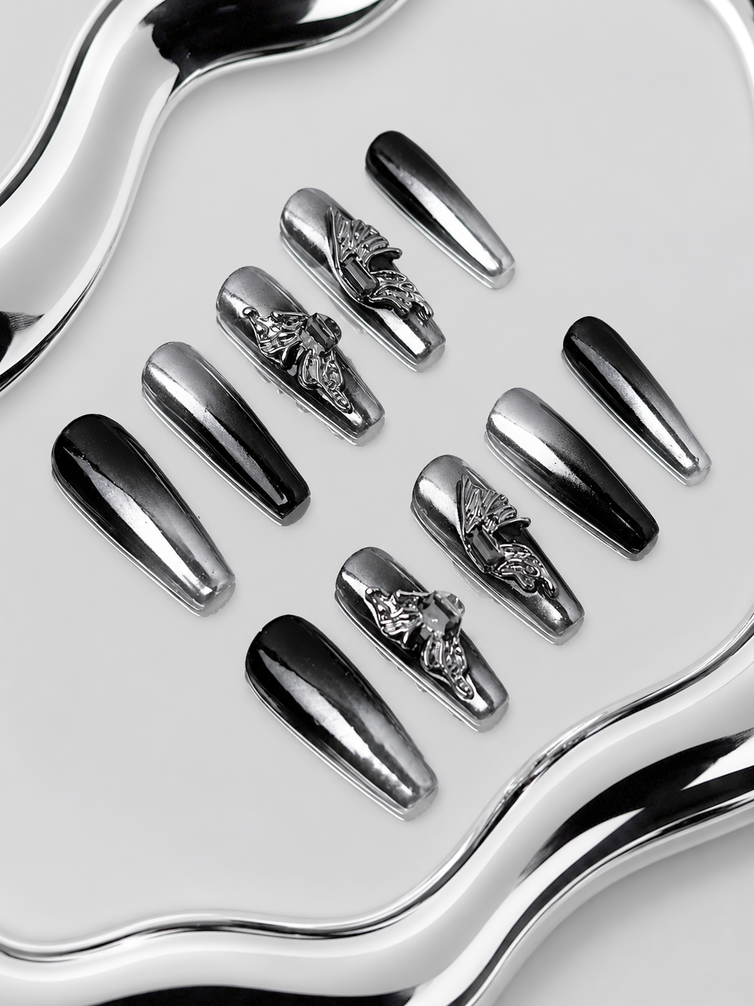Ten coffin-shaped press-on nails arranged on a reflective silver tray, featuring a mix of glossy black, metallic silver, and silver nails with raised gothic floral and scroll designs.