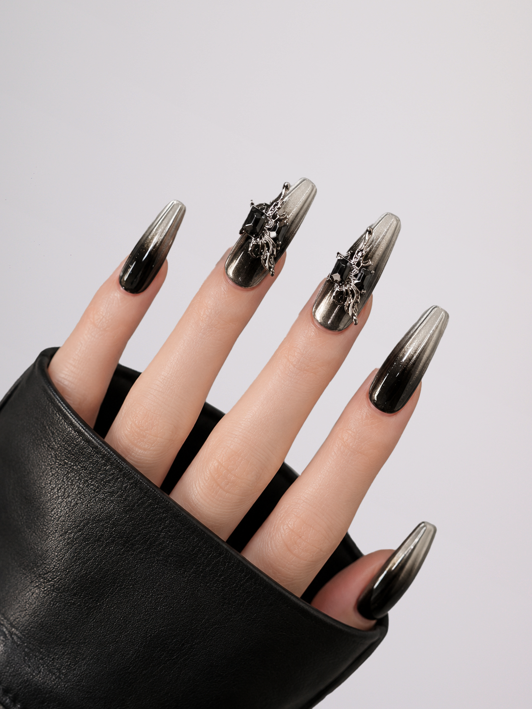 A close-up of a hand wearing long coffin-shaped nails with a black-to-silver ombre gradient and gothic silver filigree embellishments on two nails, paired with a black leather sleeve against a light gray background.