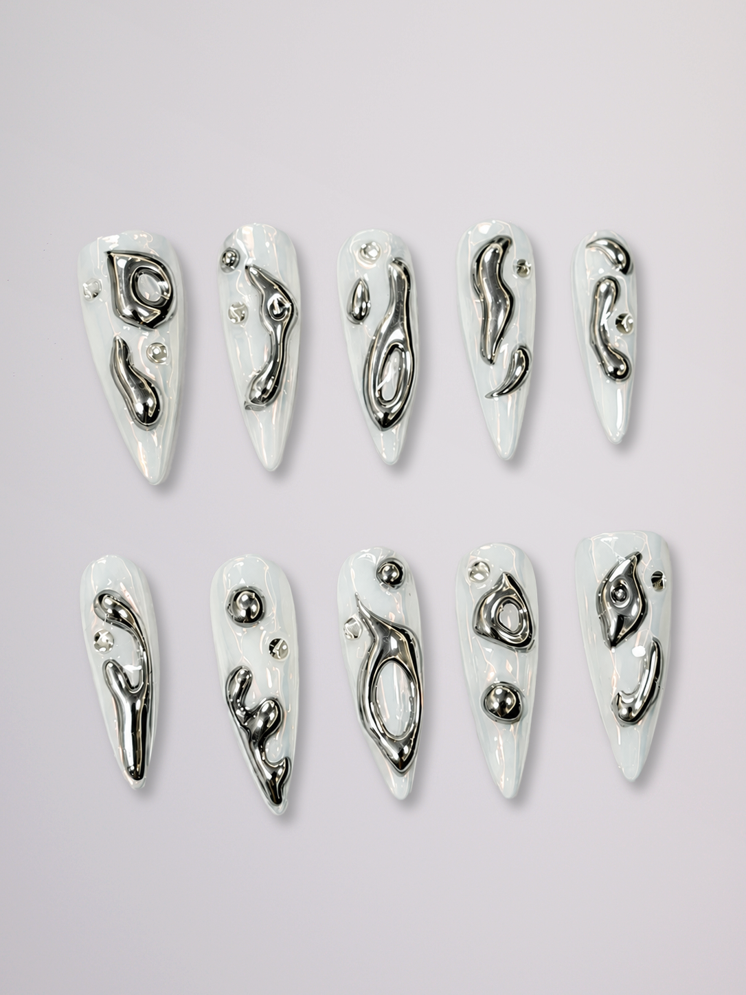 A pair of hands with long, sculpted stiletto-shaped nails decorated with silver glitter and black floral patterns, styled with multiple silver rings against a solid light gray background.