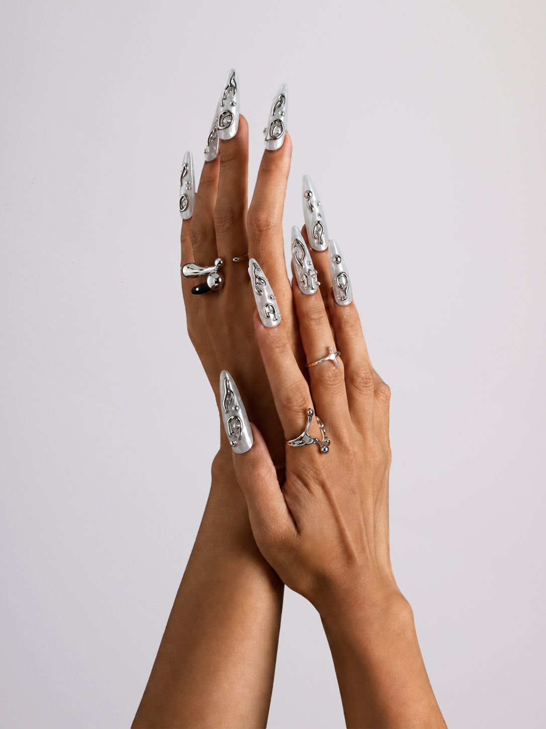 A pair of hands with long, sculpted stiletto-shaped nails decorated with silver glitter and black floral patterns, styled with multiple silver rings against a solid light gray background.