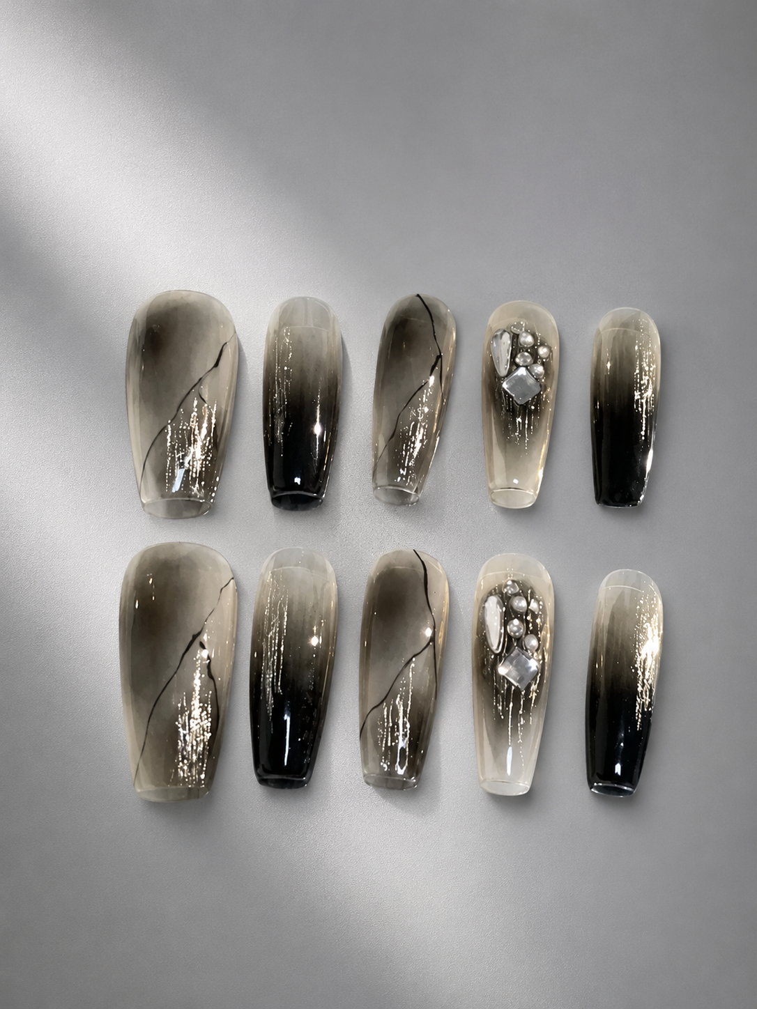 Ten false press-on nails arranged in two rows of five on a light gray background. The nails feature a smoky gradient design transitioning from translucent beige-gray at the tips to glossy black at the cuticles, with two nails decorated with silver glitter and rhinestone embellishments.