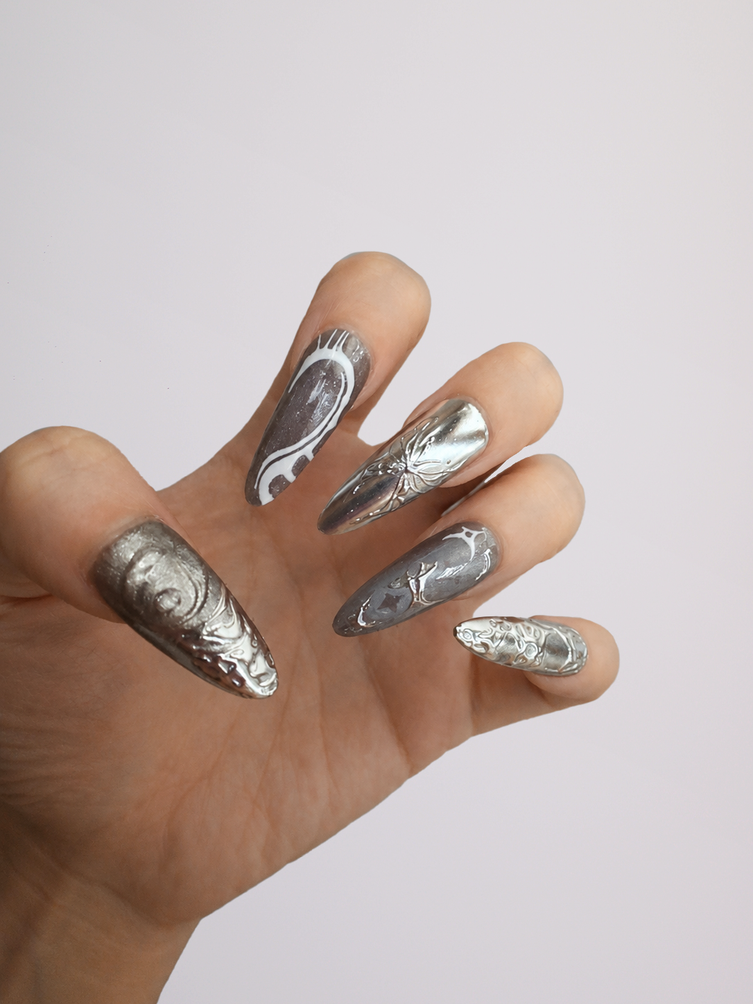 A close-up of a person's hands showcasing a silver and white stiletto nail art design with metallic rhinestone embellishments, set against a plain light gray background.