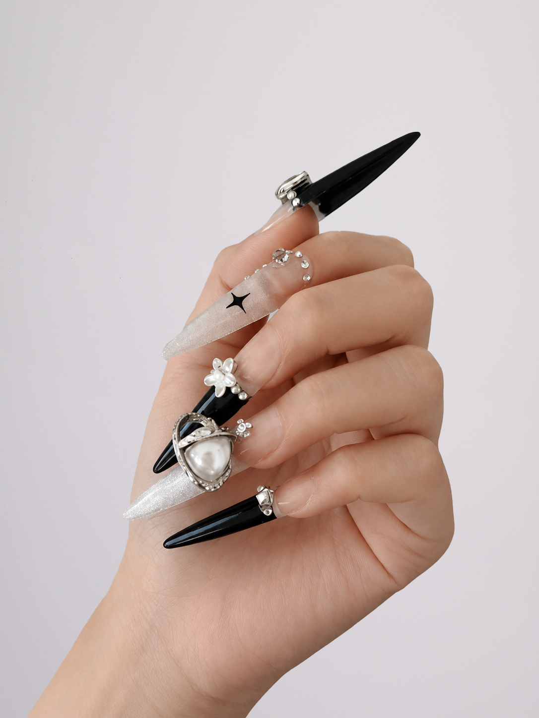 A hand showcasing a set of long, stiletto-shaped press-on nails. The nails feature a mix of glossy black and translucent white tips, decorated with silver rhinestone rings, a small black star on one nail, and a heart-and-pearl design on another.