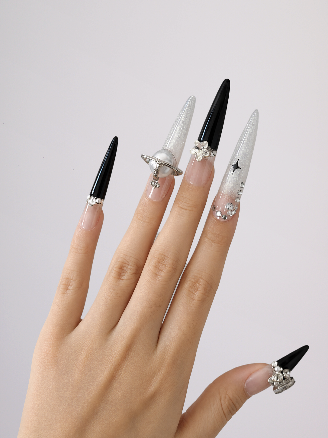 A hand displaying long stiletto-shaped press-on nails with a mix of solid black, clear glitter, and silver rhinestone embellishments against a plain light gray background.
