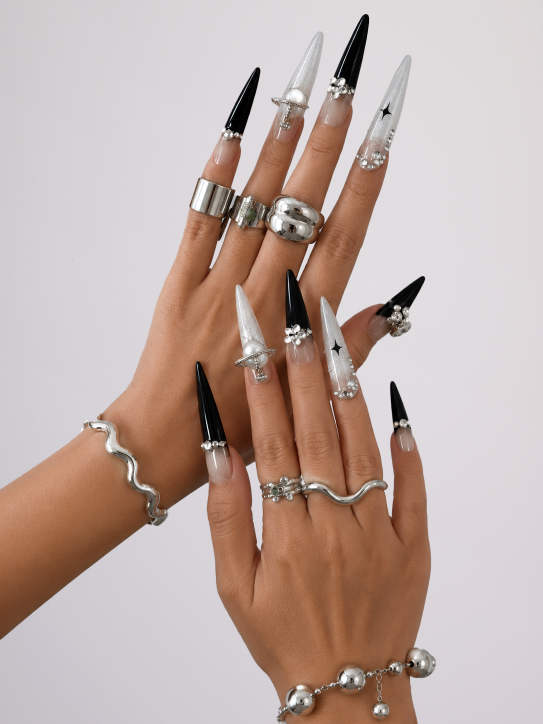 A pair of hands showcasing a gothic-inspired nail and jewelry collection. The nails are long, stiletto-shaped, alternating between glossy black and clear with silver rhinestone accents and a small black star on one nail.