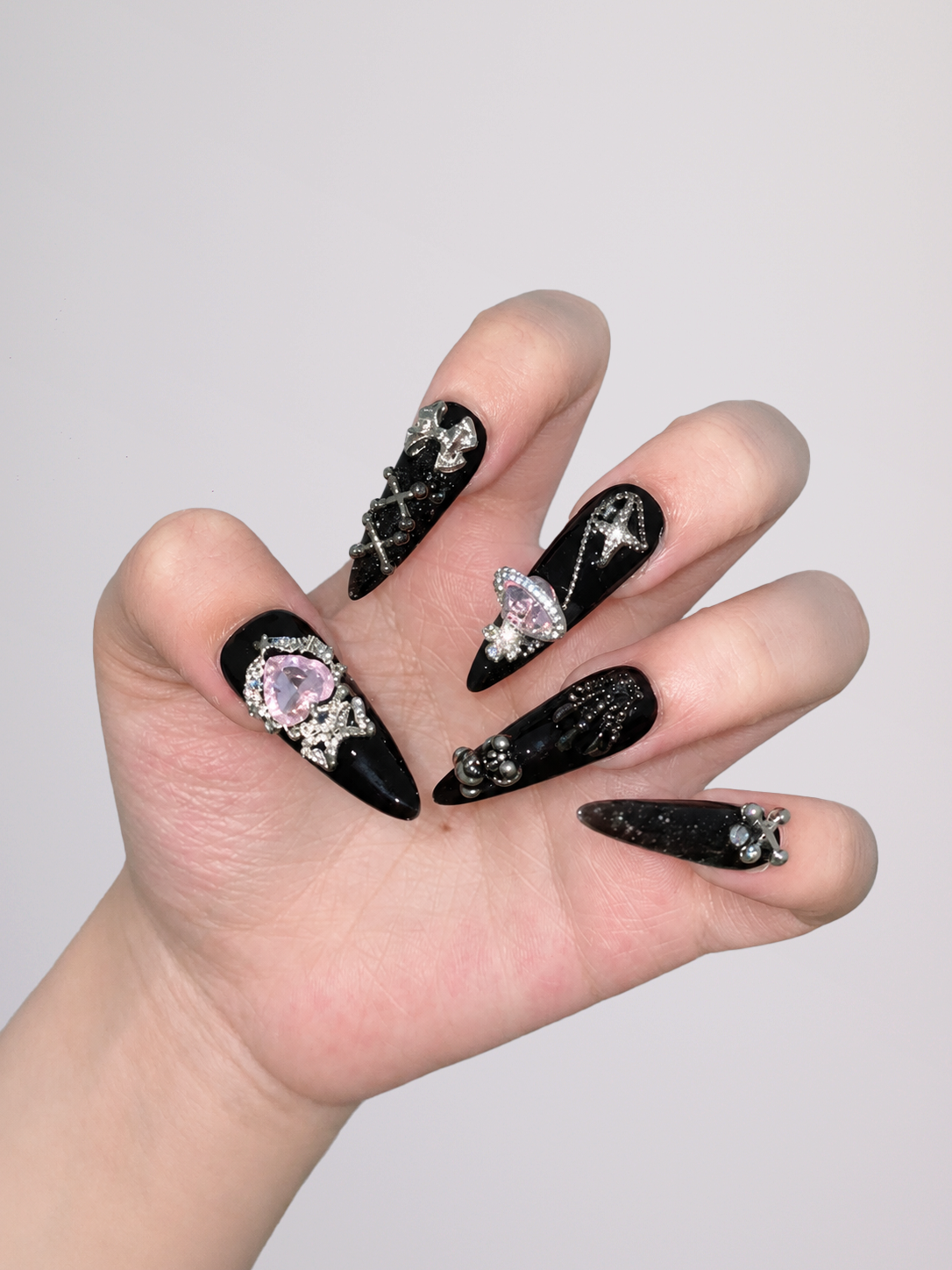 A hand with long black stiletto nails decorated with silver rhinestone chains, stars, and a pink gem on one nail, shown against a plain light gray background.