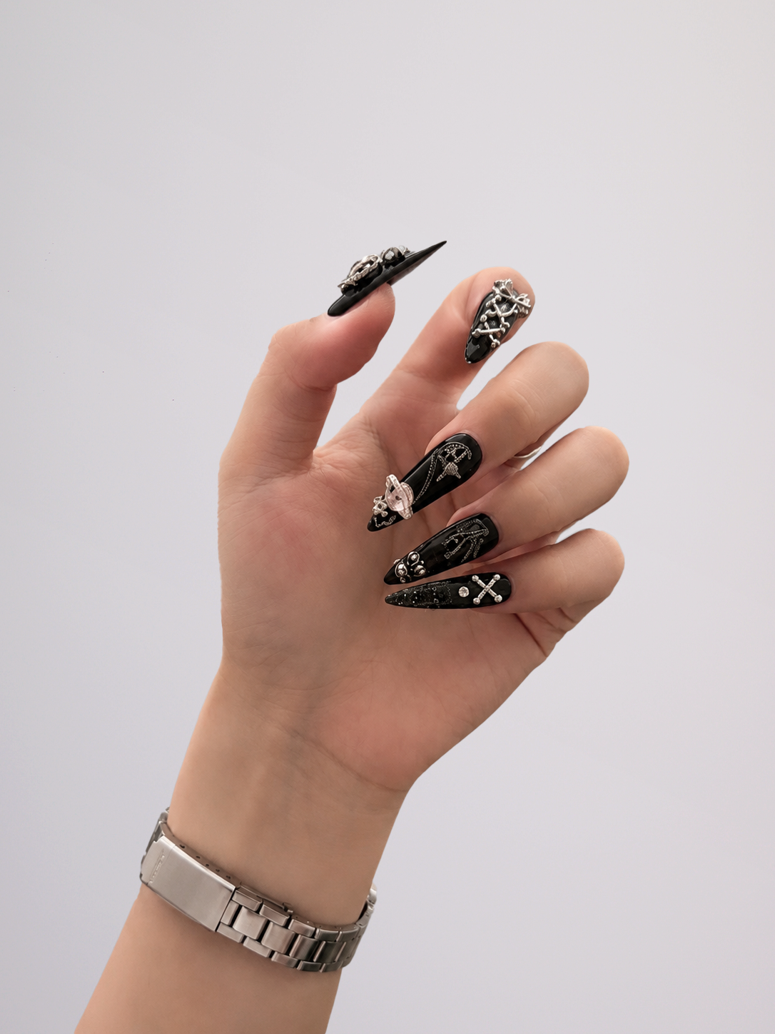 A hand with long black stiletto nails decorated with silver gothic designs, including skulls and crossbones, shown against a plain light gray background.