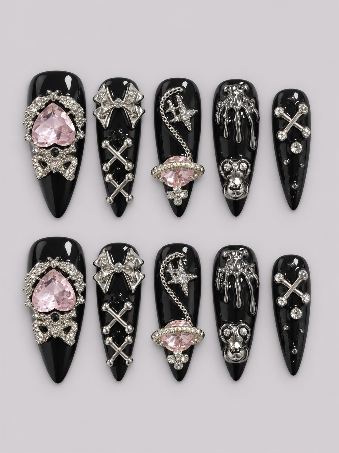 A set of 12 black coffin-shaped press-on nails arranged in two rows of six on a light gray background. The nails feature various gothic silver metal charms (bows, chains, skulls, crosses) and two pink heart-shaped rhinestone accents on different nails.