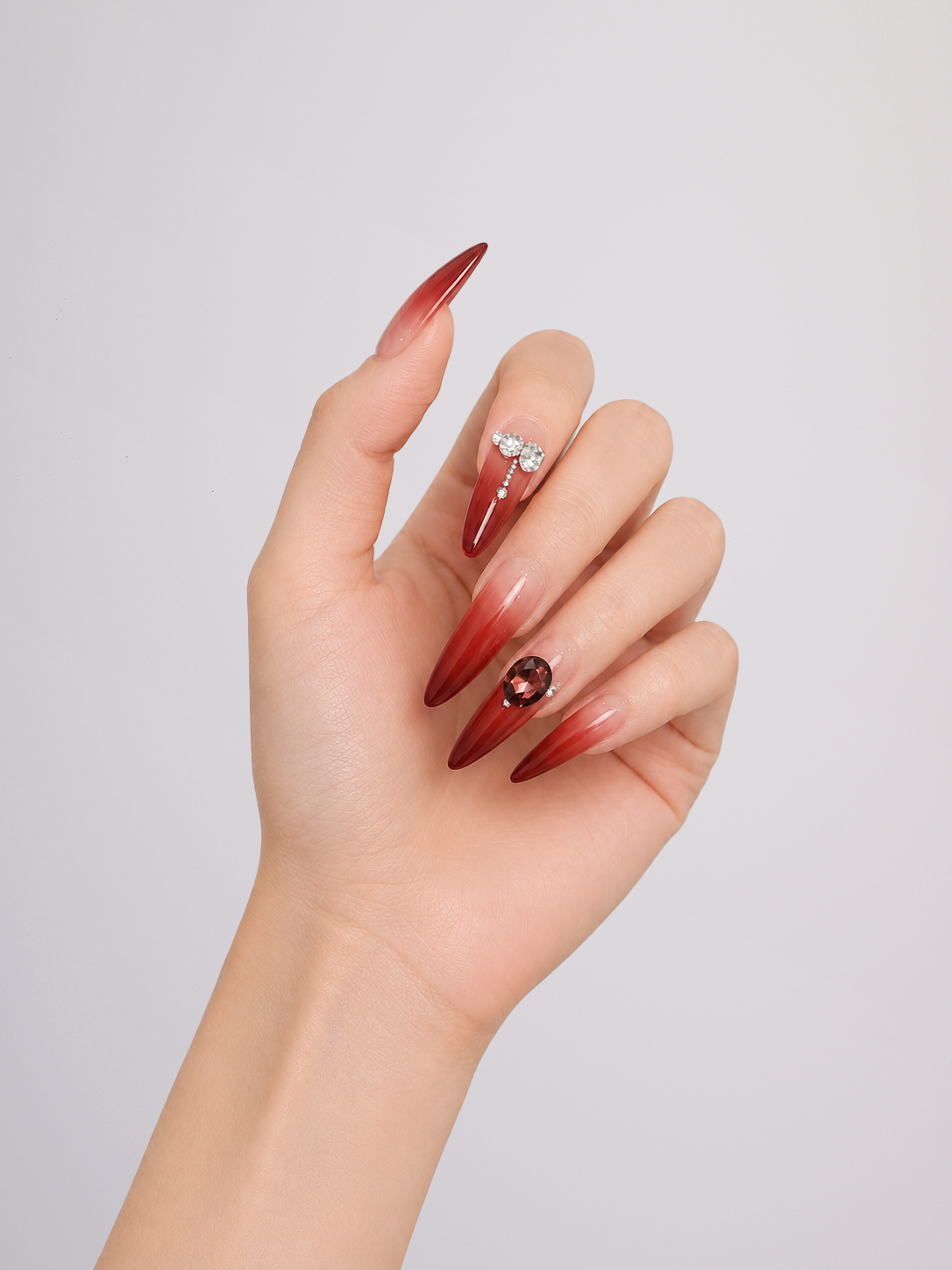 A pair of hands displaying long, stiletto-shaped press-on nails with a glossy ombre gradient from nude pink to deep burgundy, decorated with rhinestone and crystal accents.