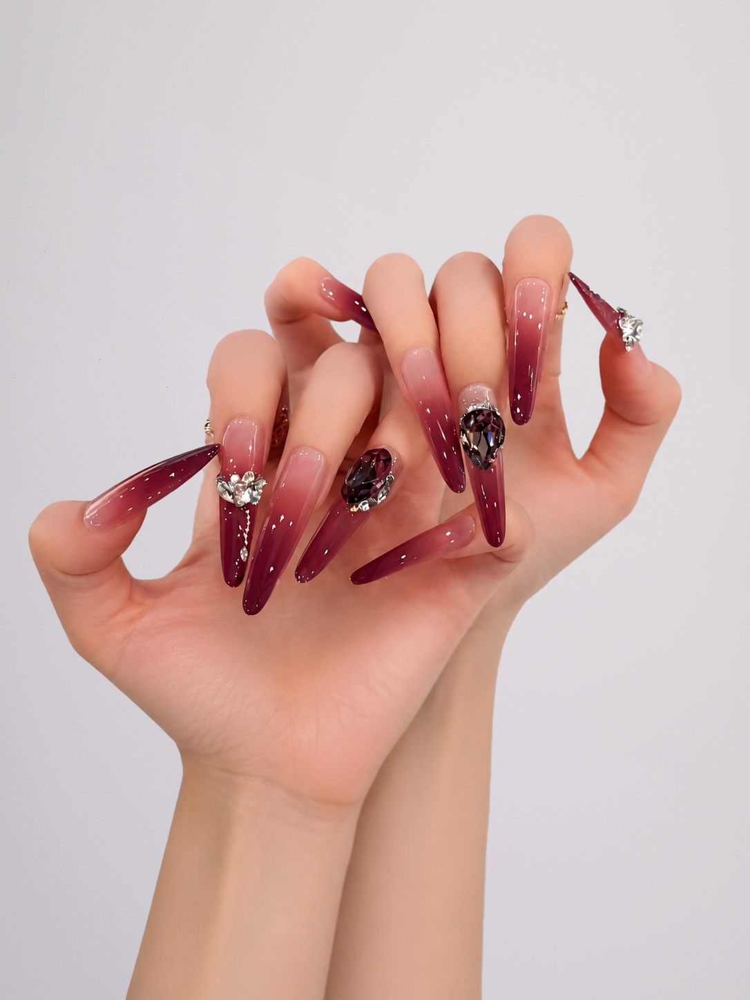 A pair of hands displaying long, stiletto-shaped press-on nails with a glossy ombre gradient from nude pink to deep burgundy, decorated with rhinestone and crystal accents.