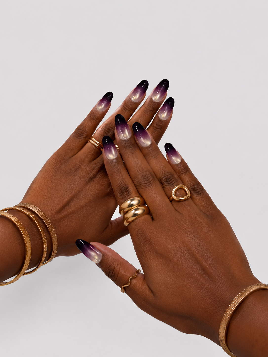 A close-up of two hands with long almond-shaped nails featuring a black-to-white-to-purple ombre gradient design, accessorized with gold rings and bracelets against a light gray background.