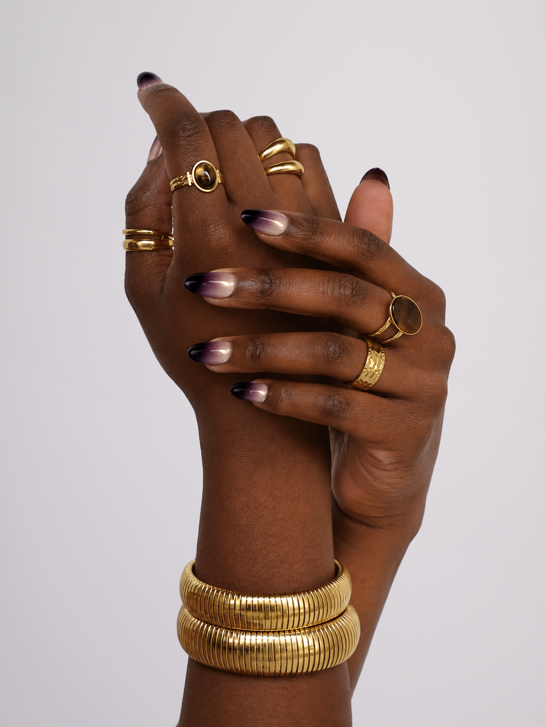 A close-up of two hands clasped together, showcasing multiple gold rings and stacked gold bangles. The nails feature a dark-to-light gradient manicure.