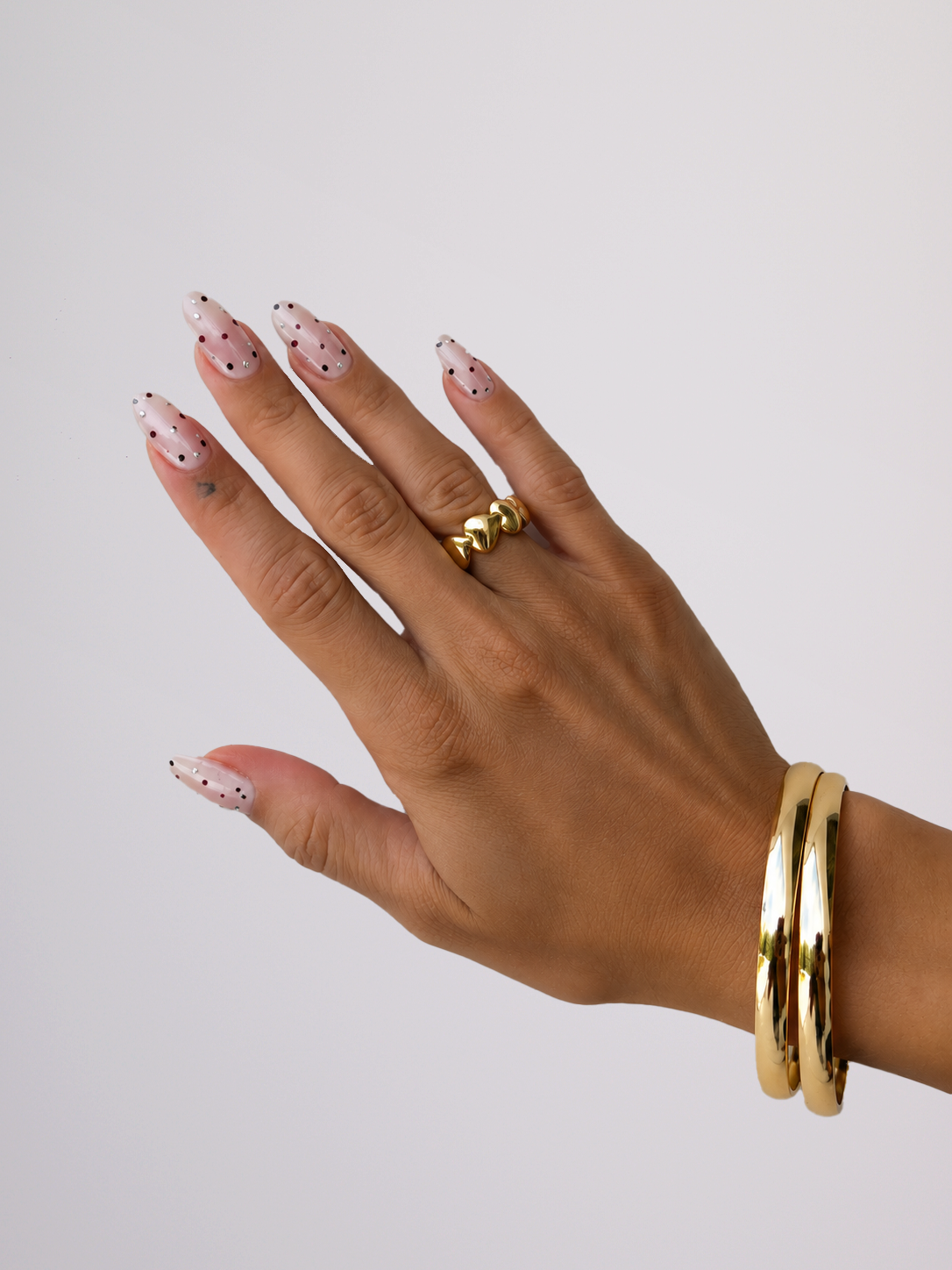 A close-up of a hand with long pink almond-shaped nails decorated with small black polka dots, wearing a chunky gold ring and stacked gold bangles against a plain light gray background.