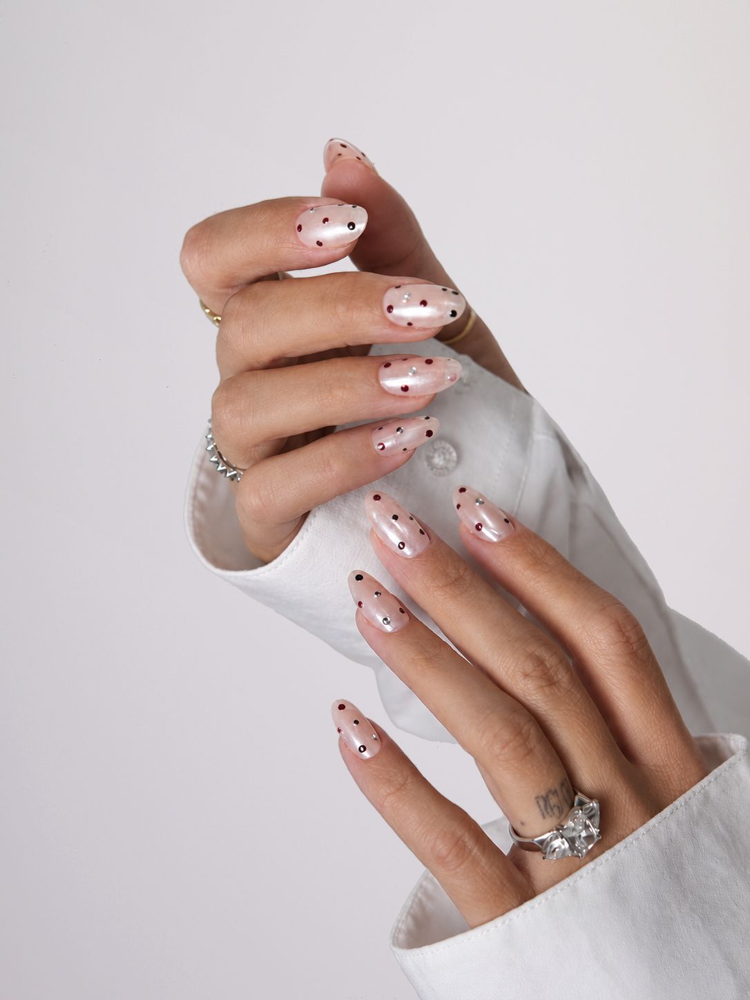 A close-up of a hand with long pink almond-shaped nails decorated with small black polka dots, wearing a chunky gold ring and stacked gold bangles against a plain light gray background.