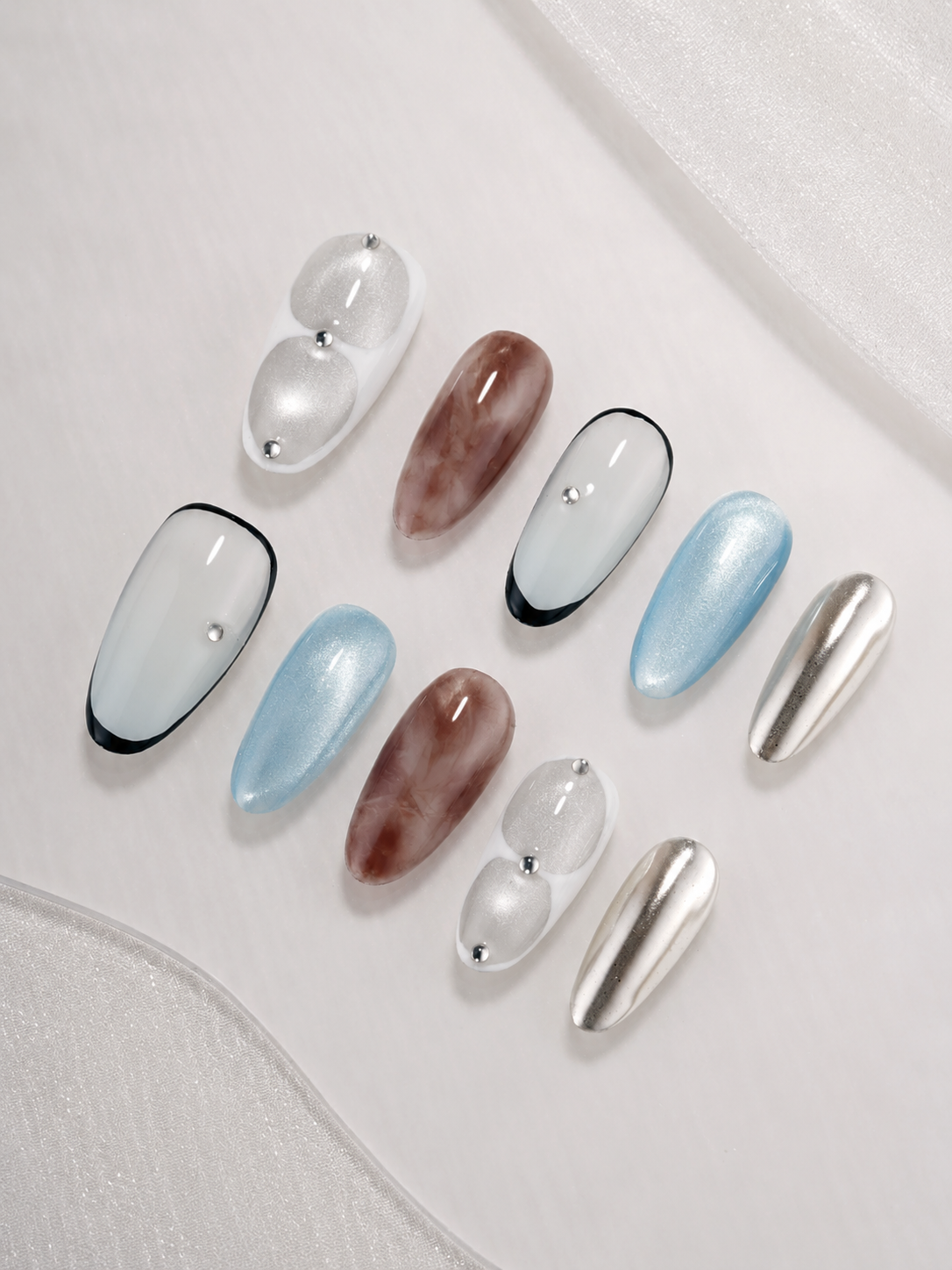 Ten press-on nails arranged on a white fabric background, featuring glossy white, light blue, brown marbled, black-framed, and metallic silver almond-shaped nails.