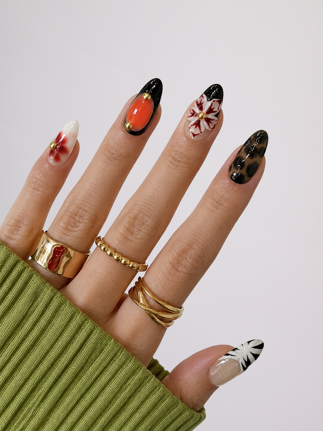 A close-up of a hand showcasing manicured almond-shaped nails with various artistic designs, including orange, black, white, and red patterns. The hand is wearing multiple gold rings and a ribbed olive green sweater sleeve, set against a clean light gray background.