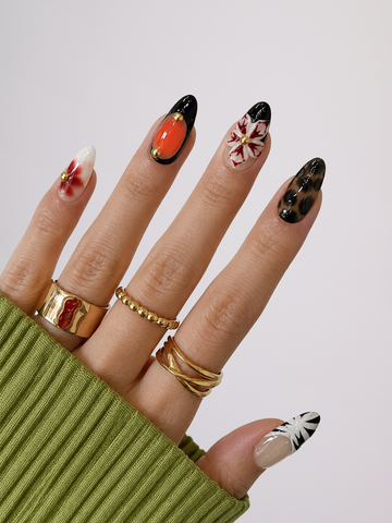 A close-up of a hand showcasing manicured almond-shaped nails with various artistic designs, including orange, black, white, and red patterns. The hand is wearing multiple gold rings and a ribbed olive green sweater sleeve, set against a clean light gray background.