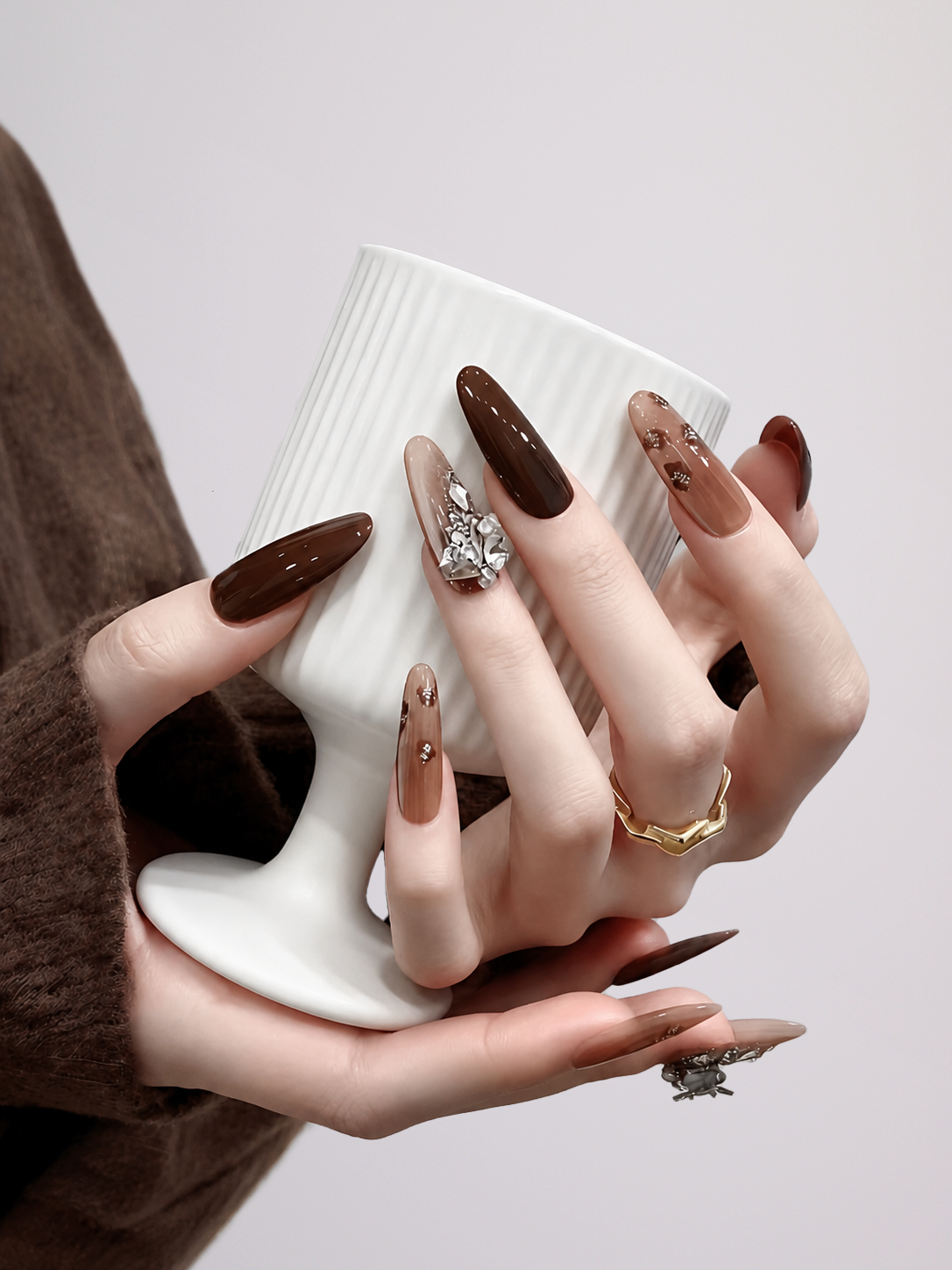 A close-up of hands with long almond-shaped brown and nude manicure holding a white ribbed ceramic cup against a light gray background.