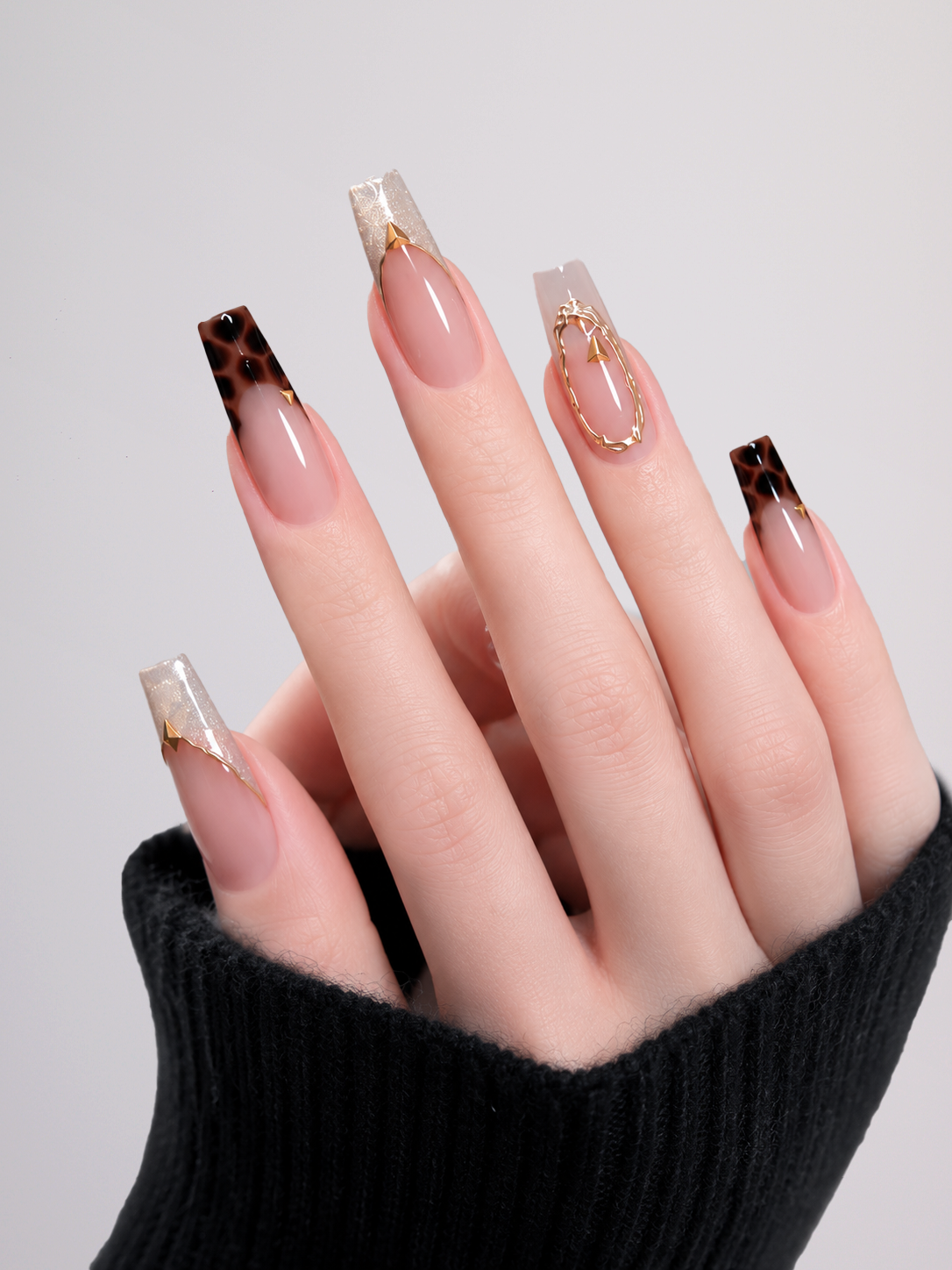 A close-up of a hand with long square press-on nails featuring a nude pink base, dark brown leopard print tips, and gold foil accents, wearing a black knit sleeve against a light gray background.
