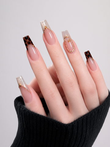 A close-up of a hand with long square press-on nails featuring a nude pink base, dark brown leopard print tips, and gold foil accents, wearing a black knit sleeve against a light gray background.