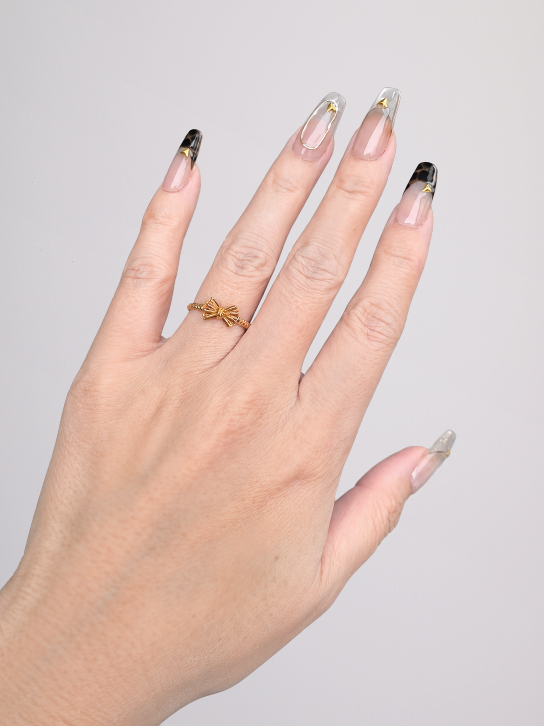 A close-up of a hand with long square press-on nails featuring a nude pink base, dark brown leopard print tips, and gold foil accents, wearing a black knit sleeve against a light gray background.