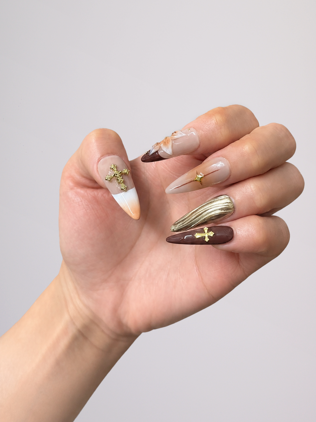 A hand displaying a set of long stiletto-shaped press-on nails with a mix of white, dark brown, and metallic gold designs, including crosses and textured patterns, against a plain light gray background.