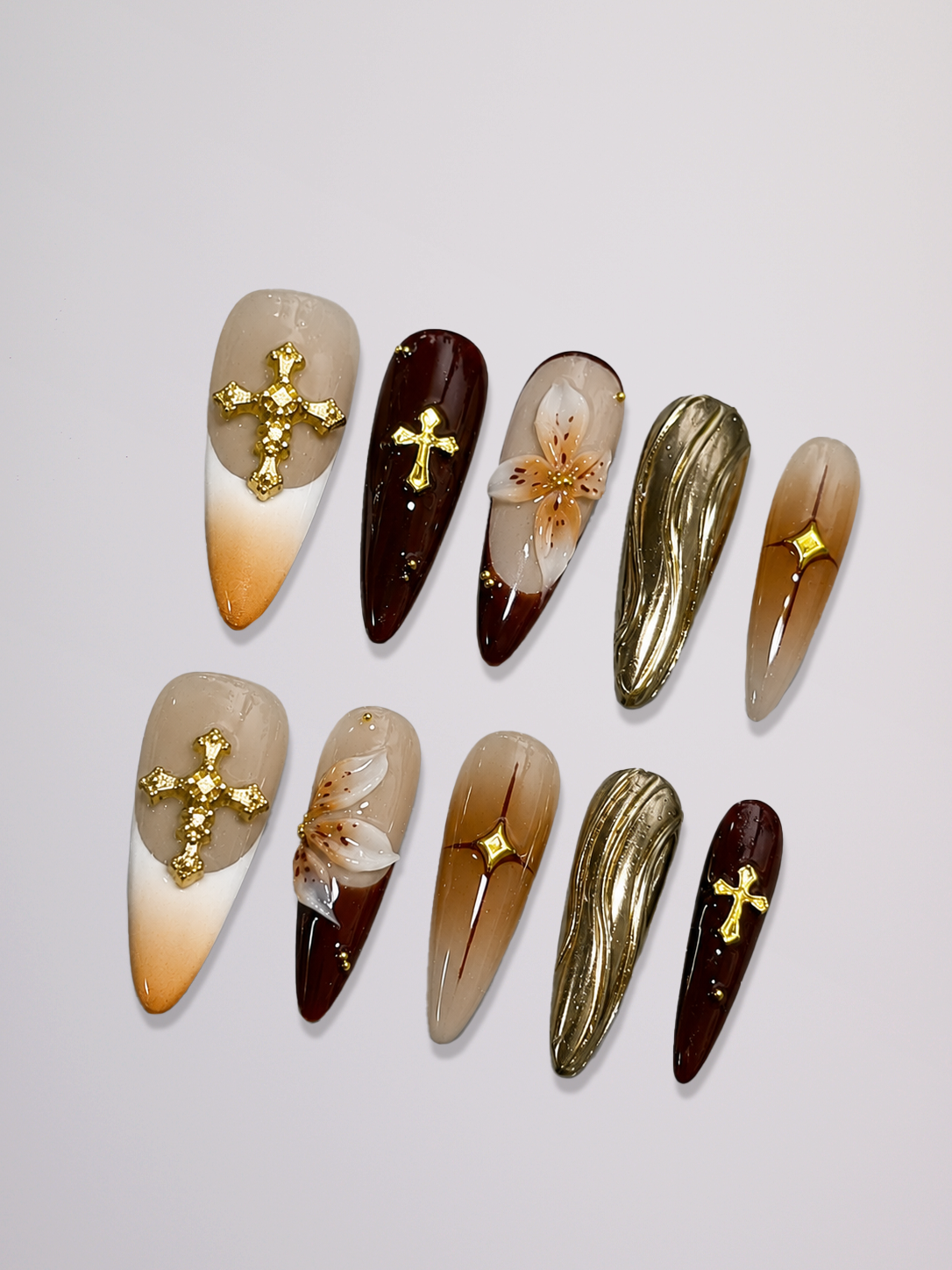 A set of ten long coffin-shaped press-on nails arranged on a light gray background. The nails feature various designs including gold crosses, white lilies, gold leaf textures, and gradient tips in shades of beige, brown, and dark brown.