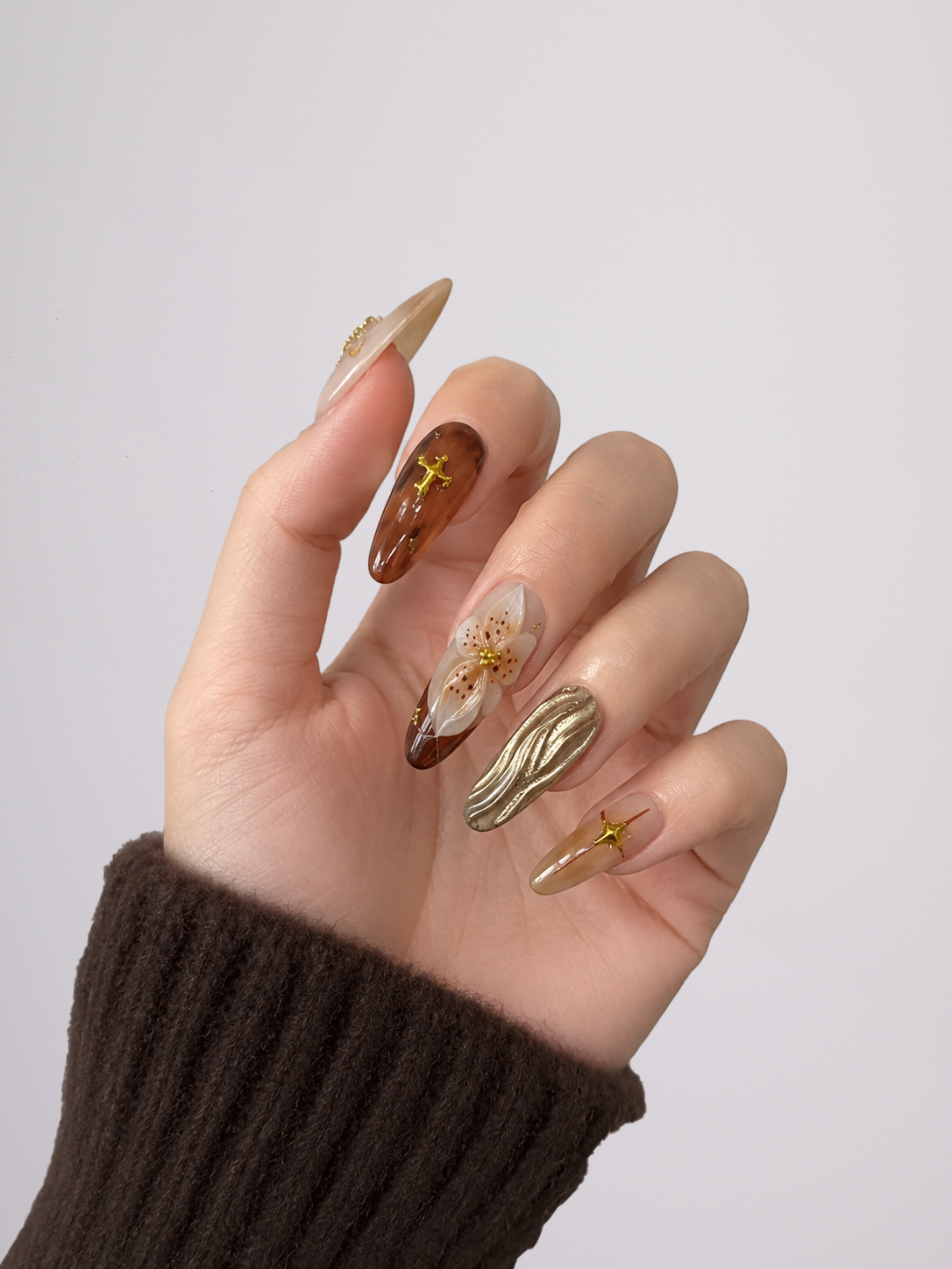 A hand with a creative, multi-design manicure featuring metallic gold, brown, and white floral/nature-inspired designs, shown against a plain light gray background.