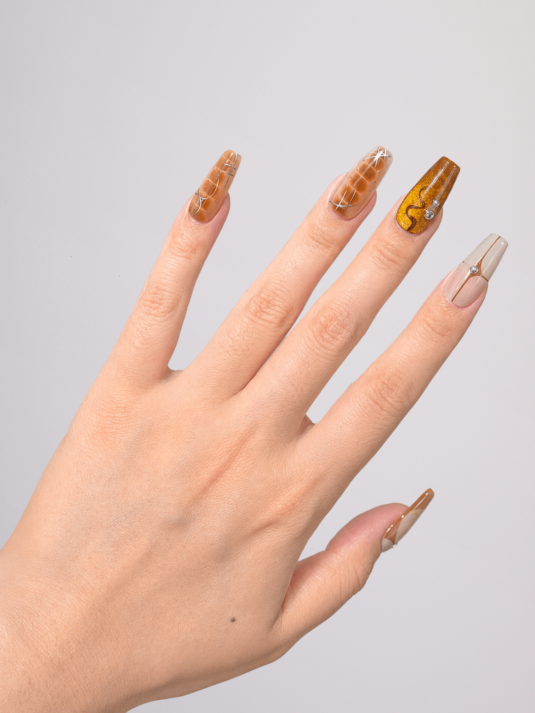 A close-up of a hand displaying long coffin-shaped press-on nails with various designs, including gold glitter, a yellow pattern, and a clear nail with a gold ring, set against a plain light gray background.