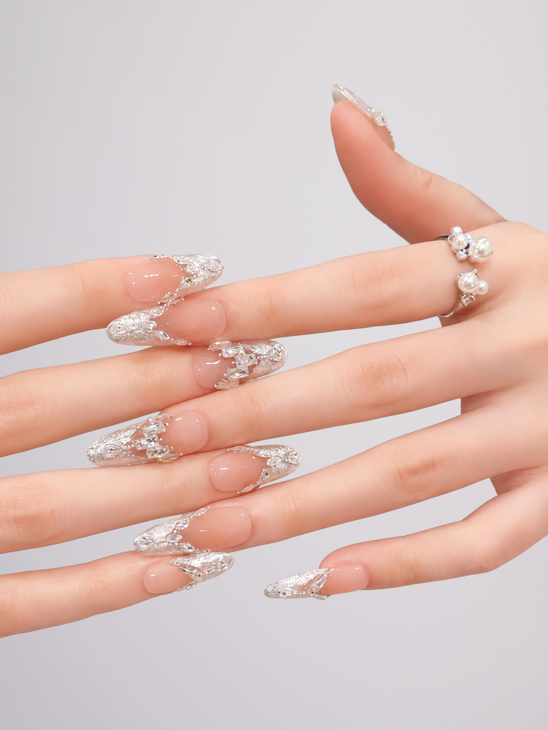 A close-up of a hand with long, sculpted stiletto-shaped nails decorated with silver rhinestone and crystal embellishments, paired with a matching silver crystal ring, set against a plain light gray background.