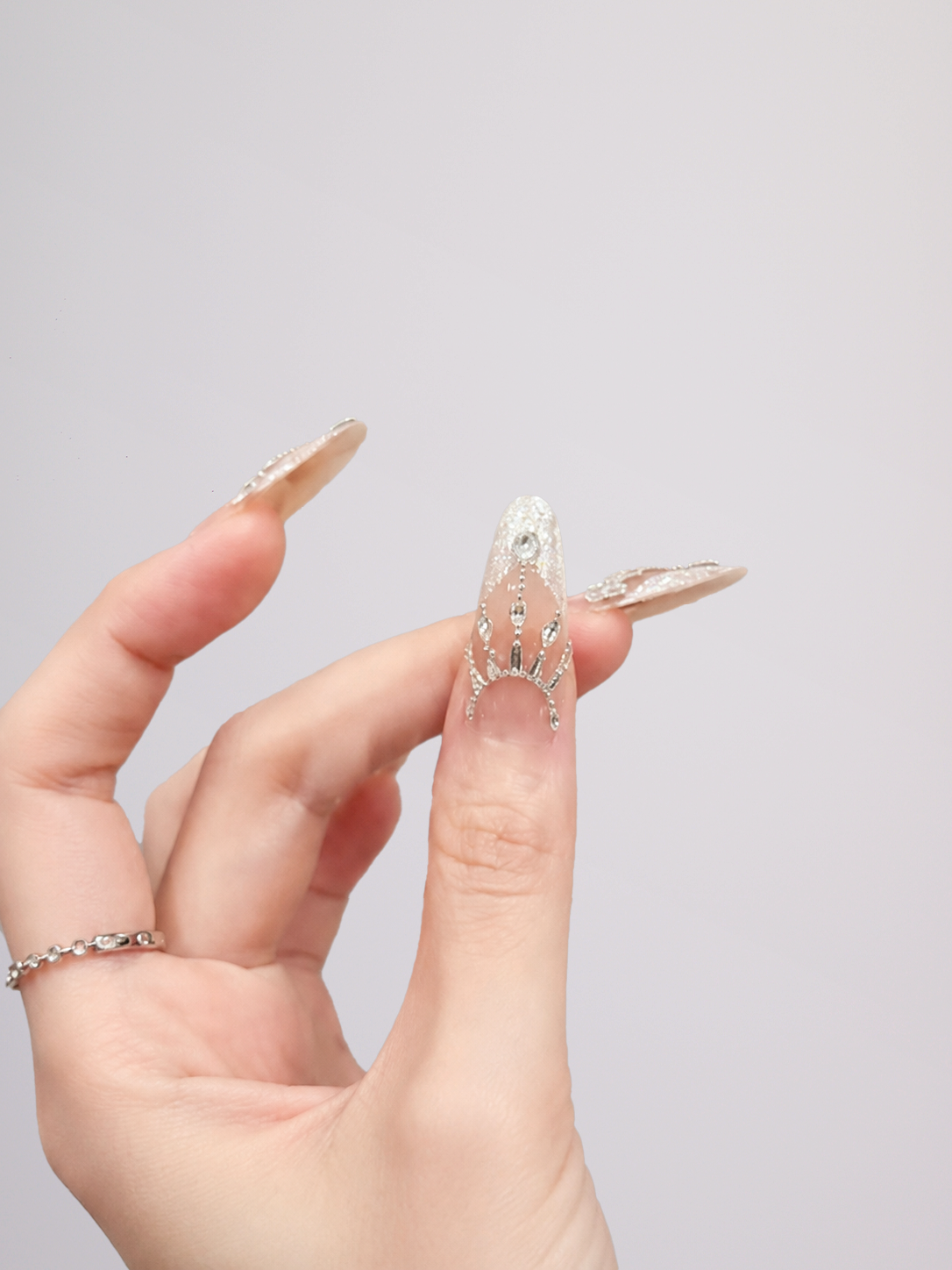 A close-up of a hand showcasing long, embellished press-on nails. The nails are clear and elongated, decorated with intricate silver rhinestone patterns and a small gold accent, with two additional clear nails featuring a gold glittery leaf design.
