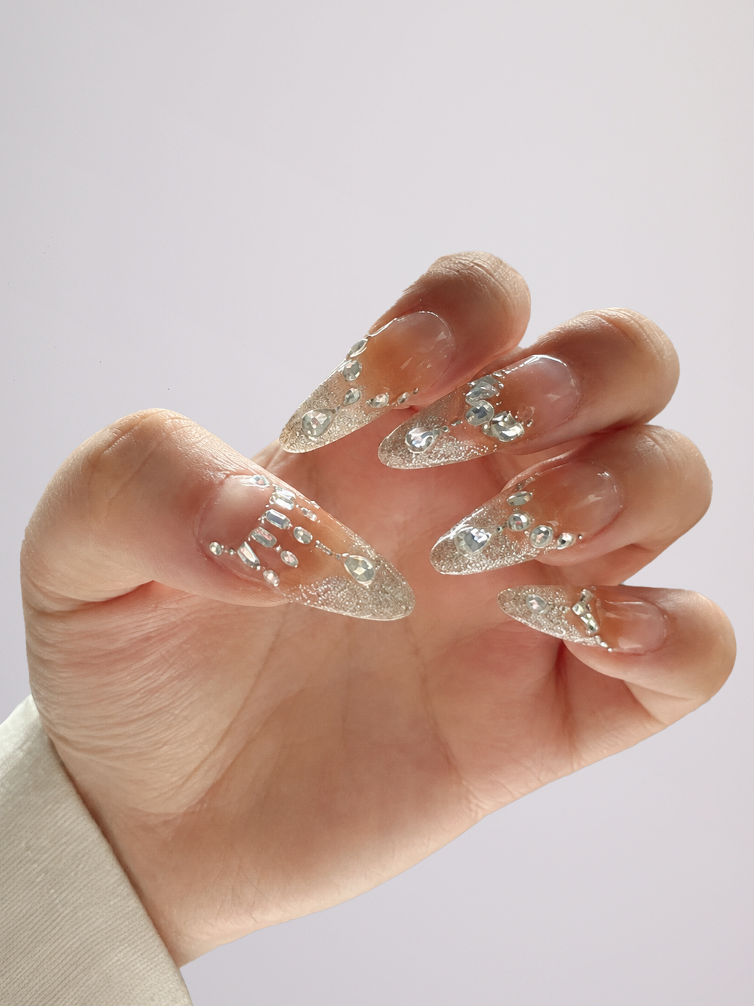 A hand with long, clear stiletto-shaped press-on nails decorated with silver rhinestones and glitter, shown against a plain light gray background.