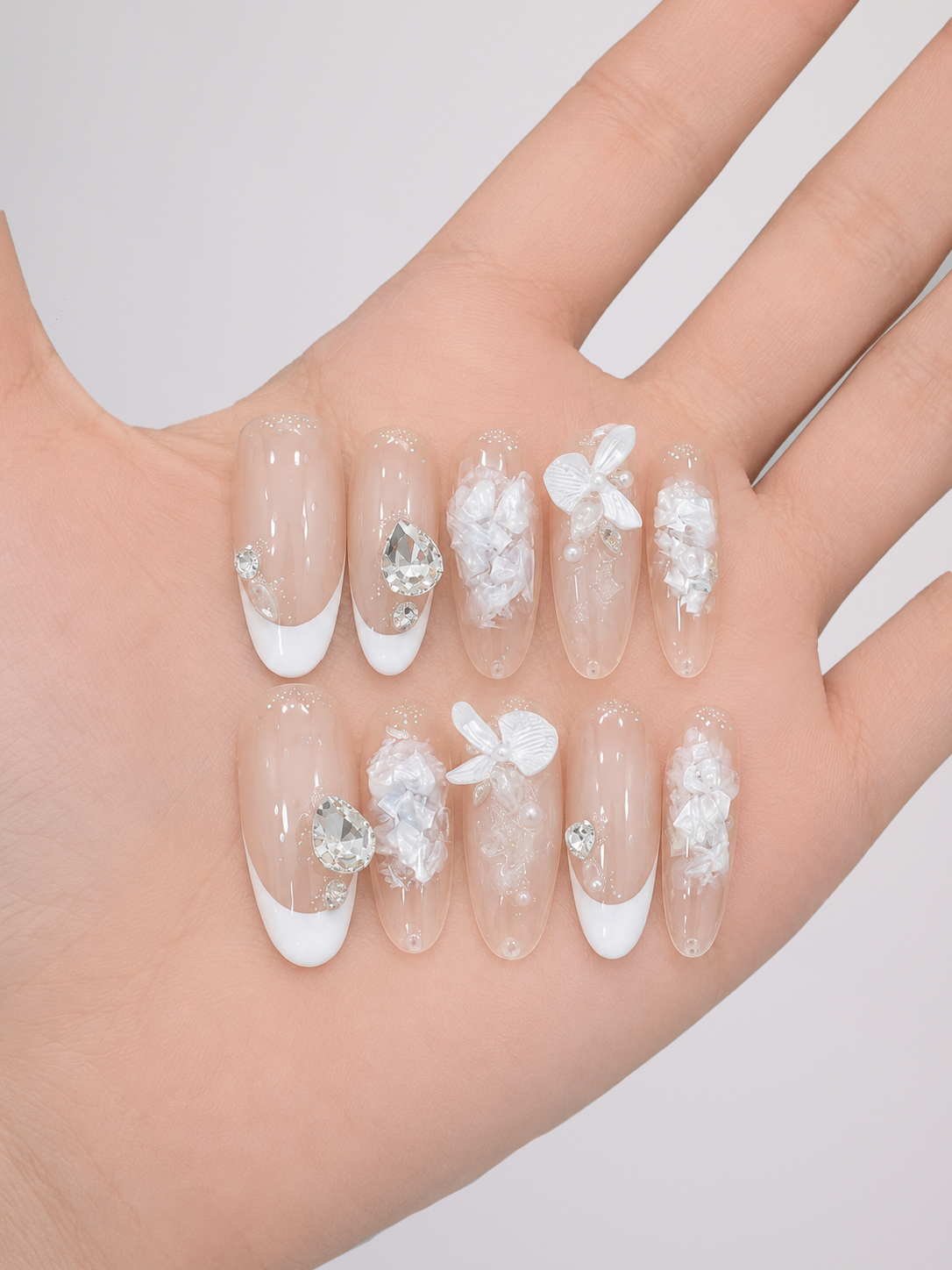 A hand displaying a set of clear press-on nails decorated with white French tips, white floral 3D elements, and silver rhinestone accents, set against a plain light gray background.