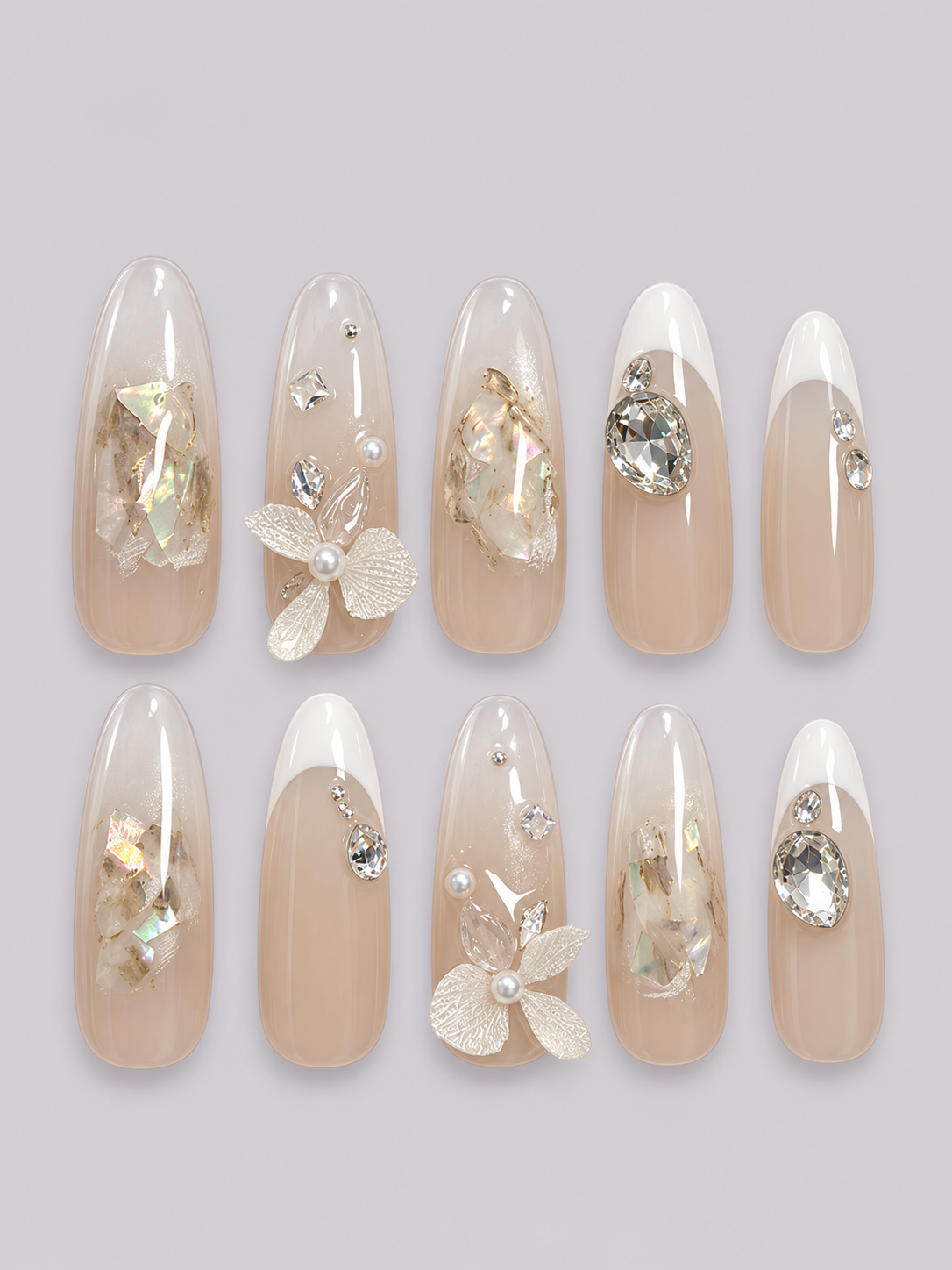 Ten almond-shaped press-on nails arranged in two rows of five on a light gray background. The nails have a glossy nude-beige base with white French tips, decorated with clear rhinestones, small white pearls, and iridescent gold foil flakes.