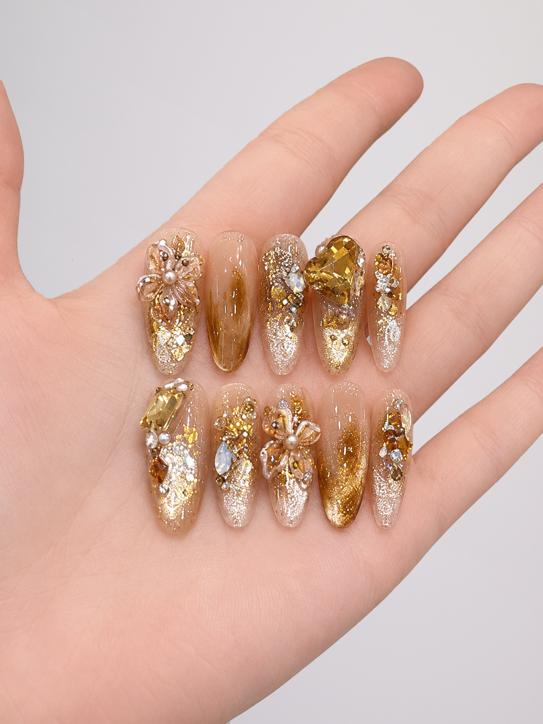 A hand displaying a set of 10 decorative press-on nails. The nails feature a mix of metallic gold glitter, clear glitter, and gold/amber gradient designs with 3D floral embellishments and small pearl accents.