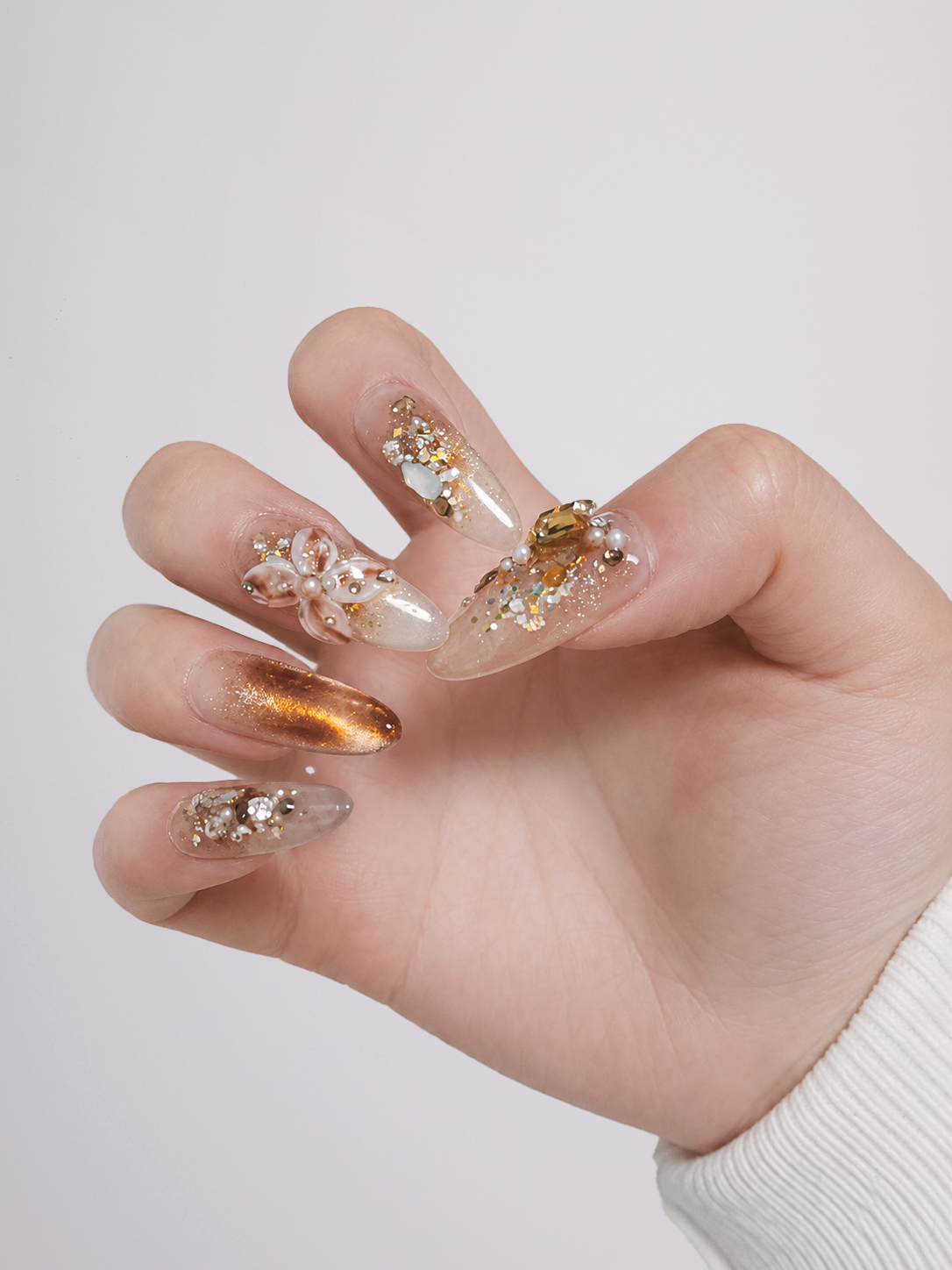A close-up of a hand showcasing long, sculpted press-on nails with a clear base, gold glitter, floral details, and rhinestone embellishments.