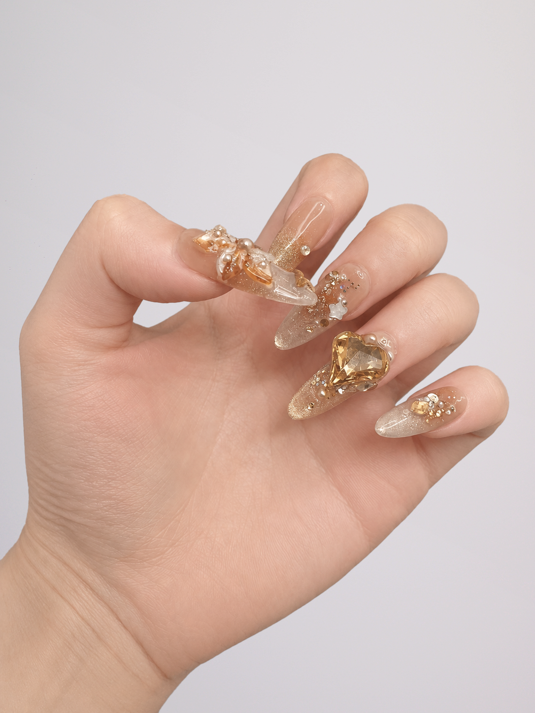 A hand with long, clear acrylic nails decorated with intricate gold glitter and metallic embellishments, shown against a plain light gray background.