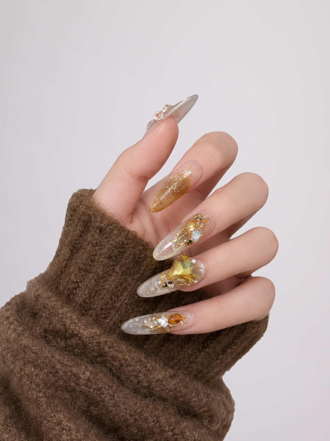 A close-up of a hand wearing long, clear press-on or manicured nails decorated with gold glitter, gold leaf, and a large gold flower on the ring finger, paired with a brown knit sleeve against a light gray background.